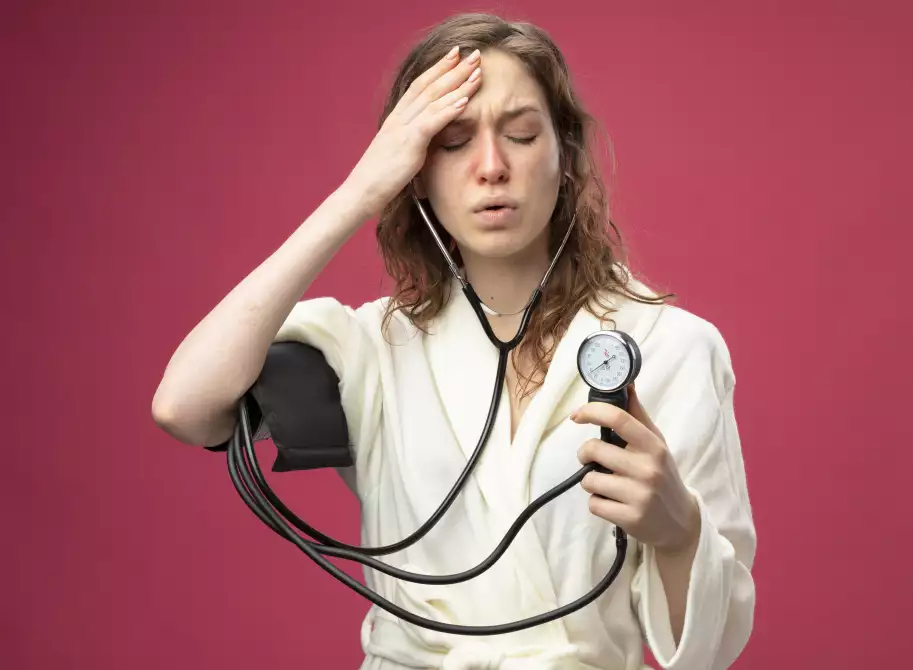 weak-young-ill-girl-with-closed-eyes-wearing-white-robe-measuring-her-own-pressure-with-sphygmomanometer-putting-hand-forehead-isolated-pink